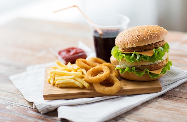 Fast Food und ungesundes Esskonzept - Nahaufnahme von Hamburger oder Cheeseburger, frittierten Tintenfischringen, Pommes Frites, Getränk und Ketchup auf Holztisch