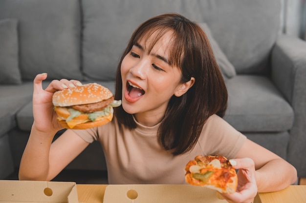 Fast Food zum Mitnehmen nach Hause. Burger essen beim Mitnehmen und Liefern. Asiatischer Frauenlebensstil im Wohnzimmer. Soziale Distanzierung und neue Normalität.