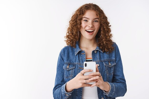 Fasziniert, gutaussehend, charismatisch, rothaariges Mädchen, lockiges Haar, sagt, Cheeze, das Sie mit der Smartphone-Kamera fotografiert, lächelt breit, ermutigt Freund, Streik, coole Pose, Foto zu machen