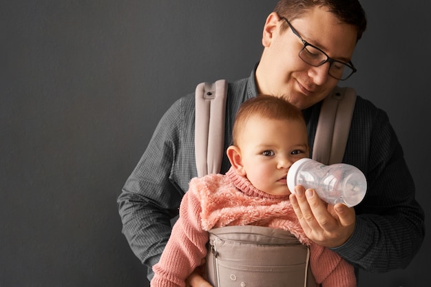 Foto fathre und sein kind in der babytrage an der grauen hintergrundwand, baby, das in der elternschaft trägt