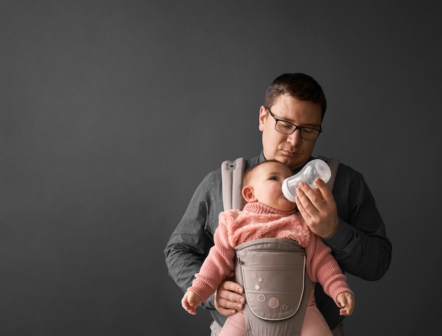 Fathre und sein Kind in der Babytrage an der grauen Hintergrundwand, Baby, das in der Elternschaft trägt