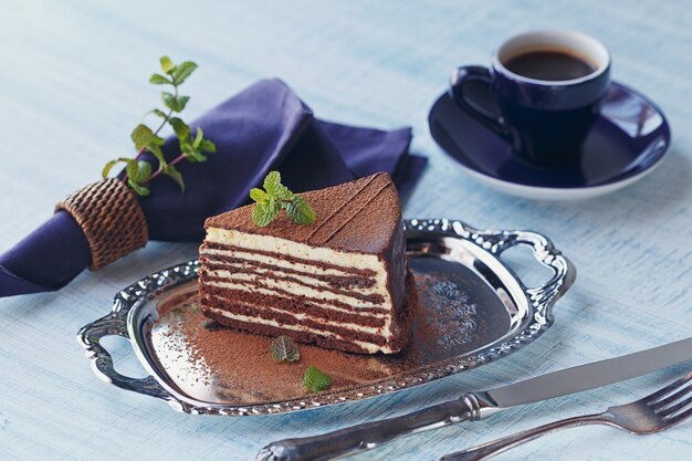 Fatia de bolo de chocolate delicioso em uma placa de prata com hortelã fresca no fundo da mesa de madeira azul claro.