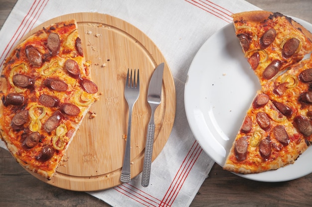 Fatia de pizza no prato, garfo, faca na mesa de madeira. Pizza no tabuleiro circular
