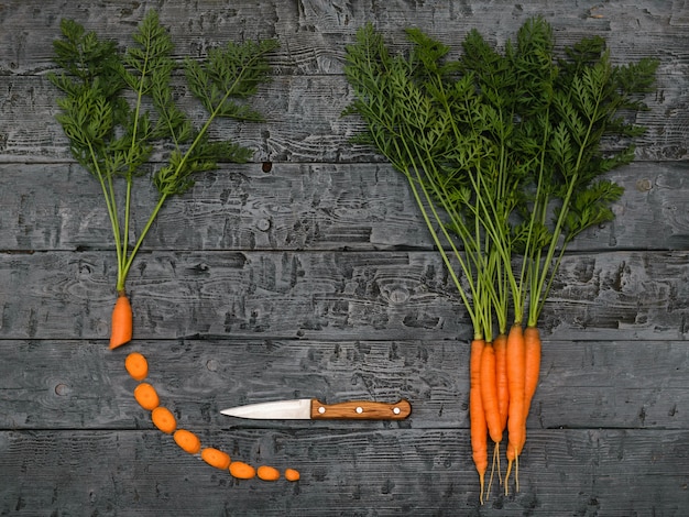 Fatias de cenoura, faca e um monte de cenouras em uma mesa rústica escura.