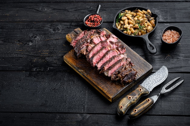 Fatias de filé de Rib Eye suculentas de carne média em uma tábua de servir de madeira com feijão branco e alecrim