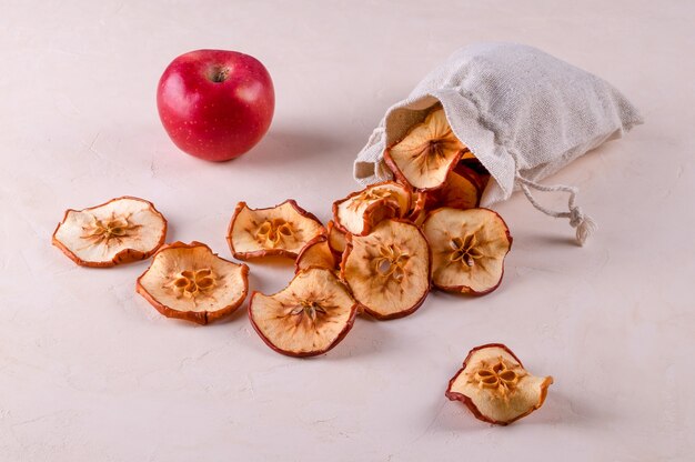 Fatias de maçãs de frutas secas em um saco ecológico e uma maçã vermelha madura repousam sobre uma superfície clara.