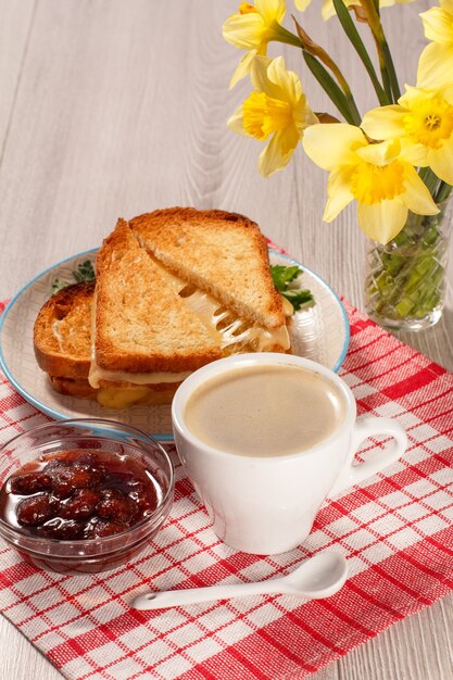 Fatias de pão torrado com queijo na chapa branca, xícara de café, colher, tigela de vidro com geléia de morango no guardanapo vermelho e buquê de narcisos amarelos. Boa comida para o café da manhã