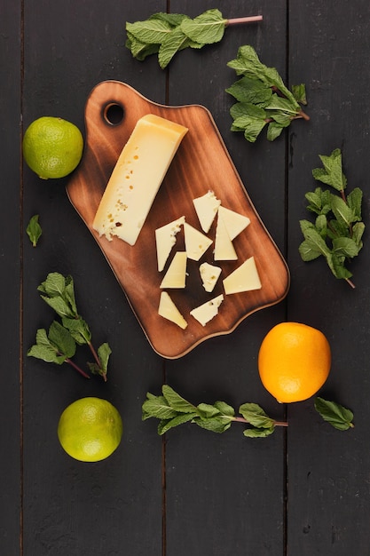 Fatias de queijo duro amarelo em uma placa de madeira com frutas cítricas com hortelã fresca em um fundo preto de madeira Fechamento de uma natureza morta Vista de cima