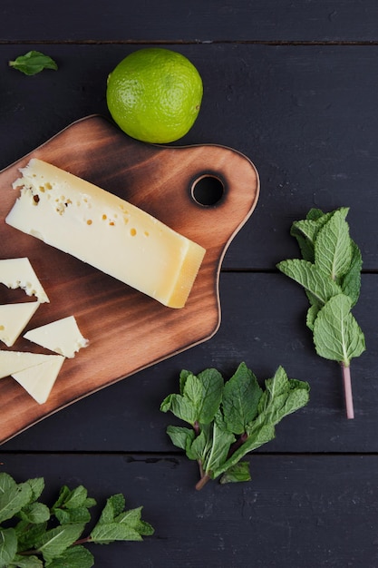 Fatias de queijo duro amarelo em uma placa de madeira com frutas cítricas com hortelã fresca em um fundo preto de madeira Fechamento de uma natureza morta Vista de cima