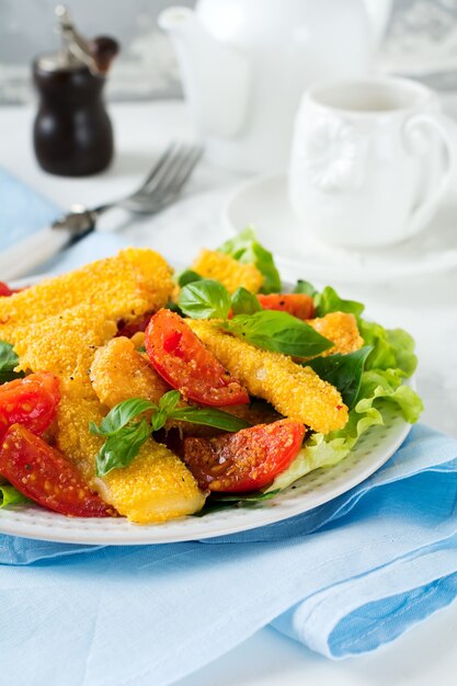 Fatias de queijo frito em grãos de milho, tomates cozidos e alface em um fundo claro. Foco seletivo.