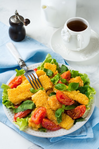 Fatias de queijo frito em grãos de milho, tomates cozidos e alface em um fundo claro. Foco seletivo.