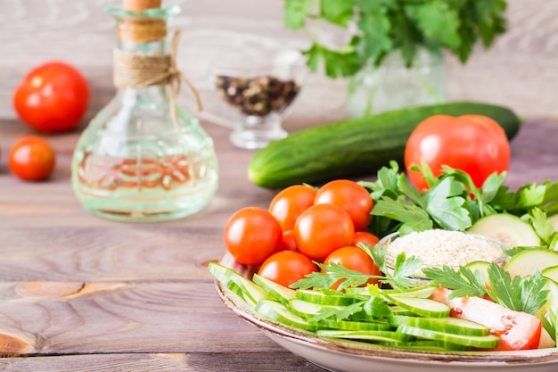 Fatias frescas de pepinos, tomates, sementes de gergelim em uma tigela e salsa folhas em um prato sobre uma mesa de madeira. Óleo e tempero para cozinhar salada de legumes