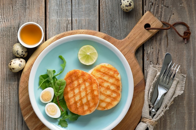 Fatias redondas grelhadas de queijo grego com mel, salada verde e ovo de codorna. Postura plana em madeira rústica