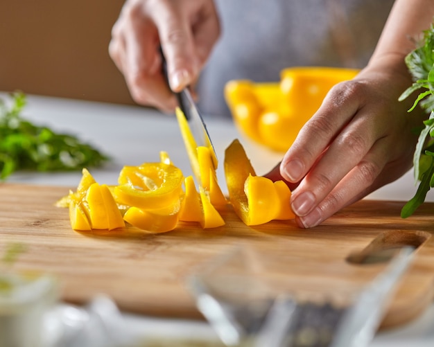 Fatias suculentas de pimenta amarela cortam as mãos de uma mulher em uma placa de madeira sobre uma mesa de cozinha branca com tomate e salsa. copie o espaço para o texto. cozinhando salada saudável passo a passo