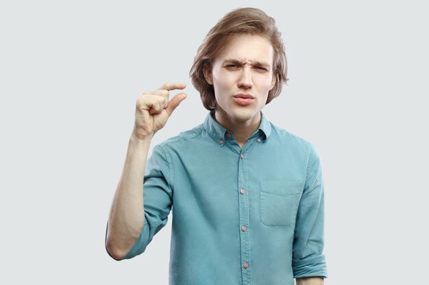 Por favor, dame algunos más. Retrato de joven rubio de pelo largo apuesto esperanzado en camisa casual azul de pie y mostrando un pequeño gesto. tiro del estudio de interior, aislado en fondo gris claro.