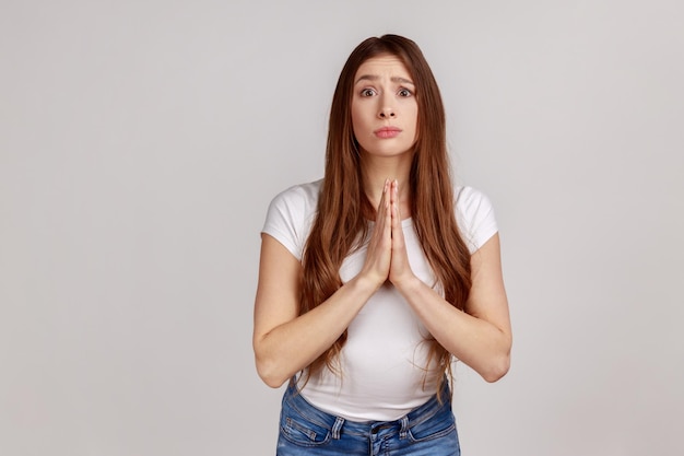 Por favor, necesito a una joven adulta tomándose de la mano en oración y pidiendo ayuda con una expresión suplicante llena de esperanza usando una camiseta blanca Foto de estudio en interiores aislada en un fondo gris