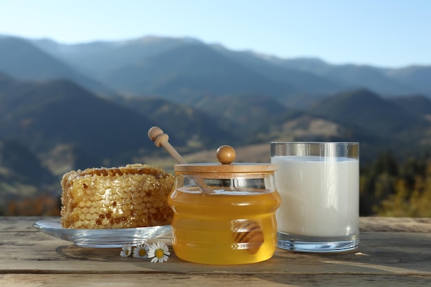 Favos de mel aromático fresco e copo de leite na mesa de madeira contra a paisagem montanhosa