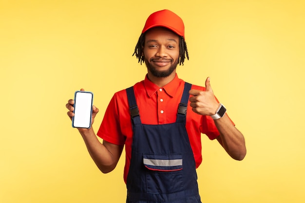 Faz-tudo satisfeito vestindo macacão segurando telefone inteligente com tela em branco para propaganda, mostrando o polegar para cima, recomendo o aplicativo on-line da indústria de serviços. Tiro de estúdio interior isolado em fundo amarelo.