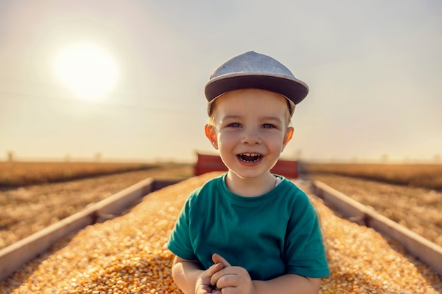 Foto fazendeiro menino bonitinho posando em um trator cheio de grãos de milho após a colheita no milharal. fazendeiro bonitinho ajudando na colheita de milho no outono. crianças e agricultura