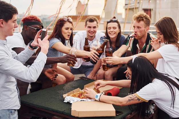 Fazendo aplausos por garrafas com cerveja Grupo de jovens em roupas casuais fazem uma festa no telhado juntos durante o dia