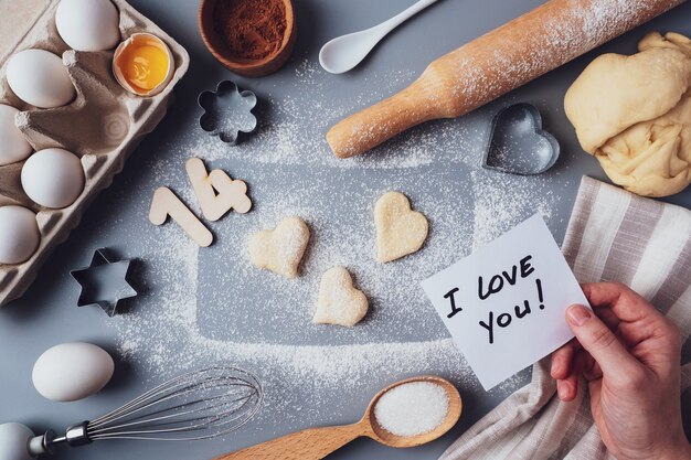 Fazendo biscoitos para o dia dos namorados, plana leiga, vista superior, copie o espaço. Vários ingredientes, rolo, massa, ovos, números 14, bata em um fundo cinza, layout em um fundo cinza.