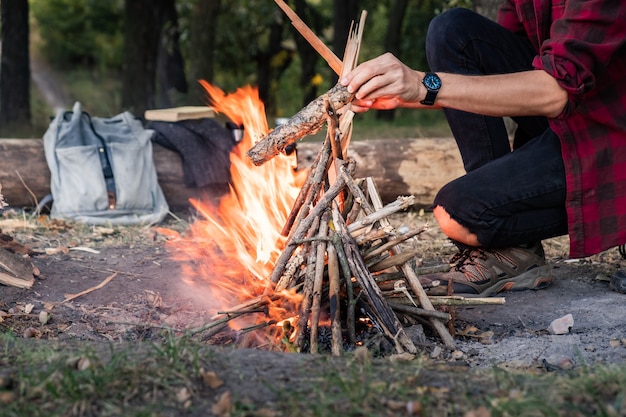 Fazendo fogueira em uma floresta. Indo para o conceito selvagem: camping com mochila vintage, garrafa térmica e roupa masculina casual coloca pedaços de lenha no fogo.