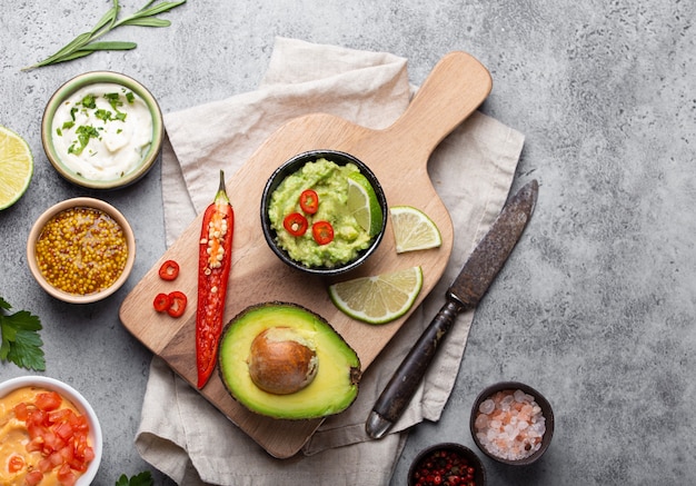 Fazendo molho de guacamole vegan verde saudável com abacate fresco, limão, pimenta na tábua de madeira com faca velha, fundo rústico de pedra, vista superior, close-up. Molho mexicano tradicional