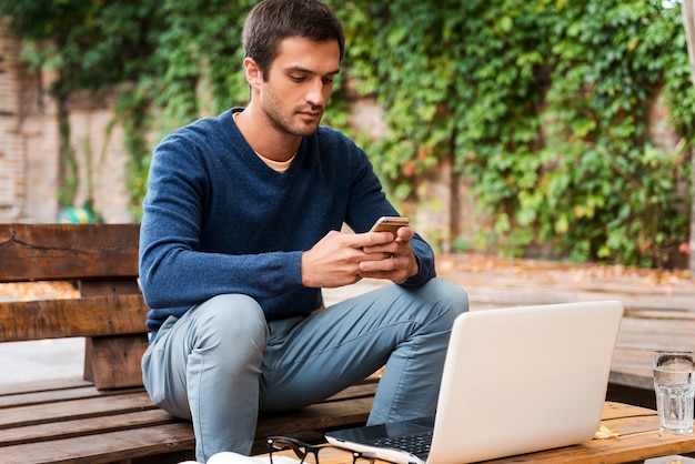 Fazendo negócios em qualquer lugar. Jovem feliz segurando um telefone celular enquanto está sentado à mesa de madeira ao ar livre