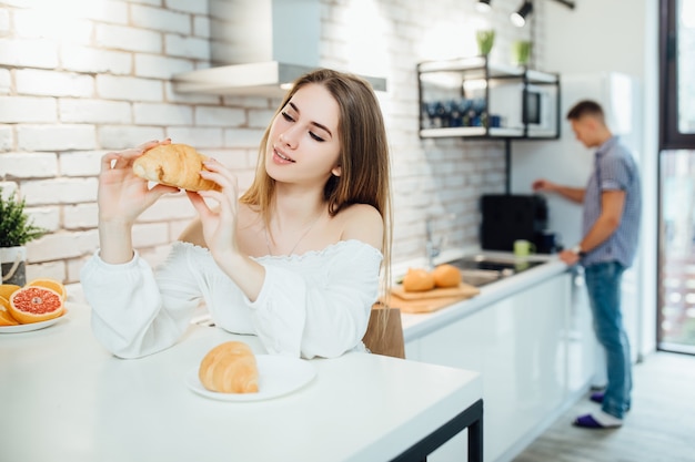 Fazendo o café da manhã na cozinha, mulher loira muito sorridente segurando um croissant, comida saudável, enquanto está sentado na cadeira.