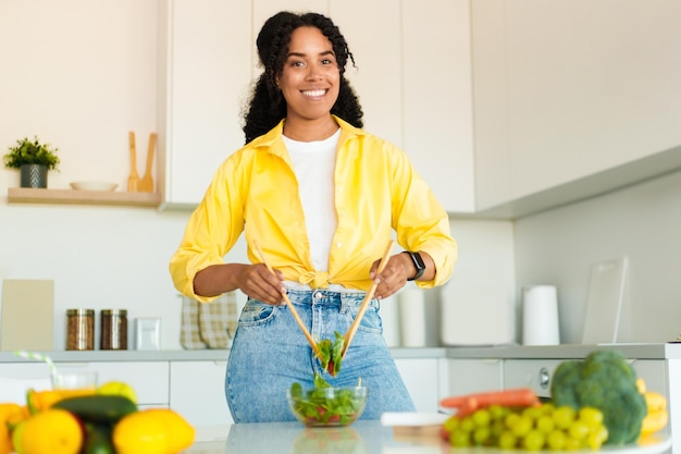 Fazendo uma refeição saudável Mulher afro-americana positiva cozinhando salada fresca saudável e sorrindo para a câmera na cozinha