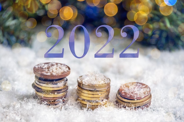 Fecha 2022 en color de moda MUY PERI. Pirámides de oro de monedas cubiertas de nieve sobre un fondo de ramas de abeto verde y bokeh de una guirnalda. Concepto de riqueza en el nuevo año.