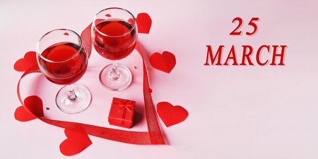 Fecha del calendario sobre fondo claro con dos copas de vino tinto, caja de regalo roja y corazones rojos 25 de marzo