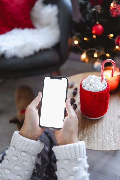 Fechar smartphone com maquete de tela vazia em plano de fundo de férias com árvore de Natal, cliente segurando o telefone, fazer compras online, comprar presentes, pesquisar informações, navegar em aplicativos