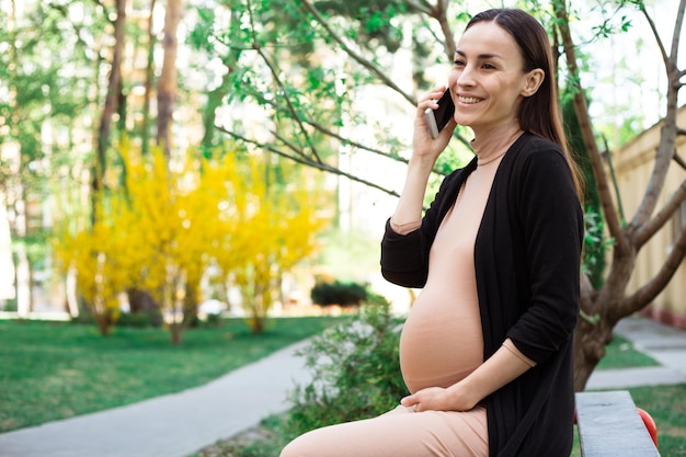 Feche a foto da mulher sorridente grávida no banco no parque enquanto ela fala no telefone inteligente