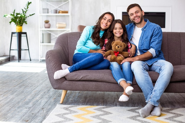 Feche a foto de mãe, pai e filha posando juntos para uma foto conjunta em sua sala de estar estilosa, se abraçando com sorrisos radiantes