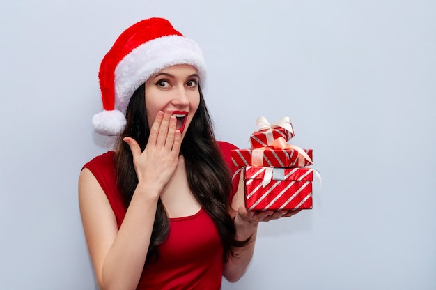 Feche a foto de Natal da garota impressionada gritando Wow OMG com caixas de presente no vestido vermelho e chapéu de Papai Noel.