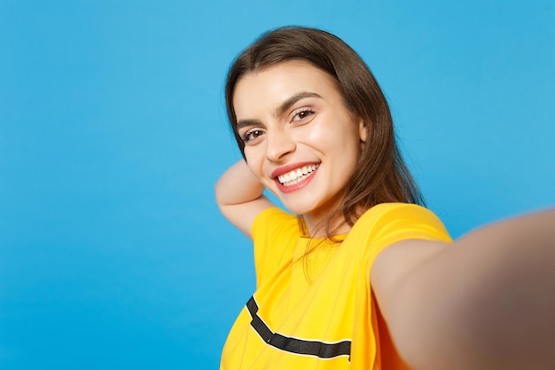 Feche a foto de selfie de uma bela jovem atraente sorridente em roupas casuais vívidas, olhando a câmera isolada no fundo da parede azul brilhante no estúdio. Conceito de estilo de vida de pessoas. Mock up espaço de cópia.