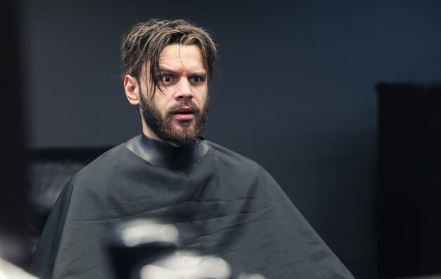 Feche a foto de um homem barbudo com cabelo bagunçado que está olhando seu reflexo no espelho de uma barbearia com uma expressão facial de medo.