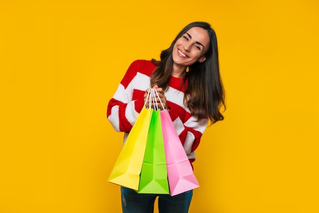 Feche a foto de uma bela jovem feliz e animada com um suéter listrado com muitas sacolas de compras coloridas e cartão de crédito nas mãos, isolado em fundo amarelo