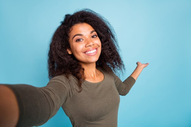 Feche a foto de uma garota positiva alegre turista afro-americana, segure a mão, convide, mostre suas férias, tome uma selfie, videochamada, use uma camisa estilo casual isolada sobre a parede azul