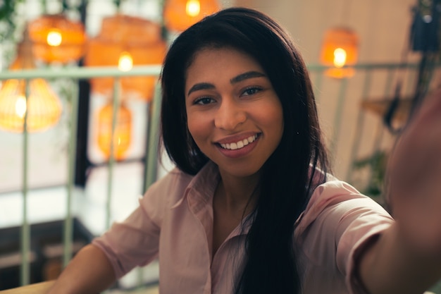 Feche a foto de uma jovem alegre, sorrindo para a câmera enquanto tira uma selfie. Lâmpadas no fundo