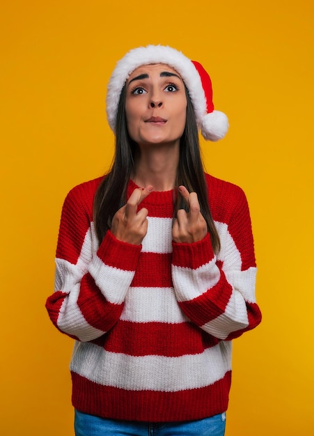 Feche a foto de uma jovem encantadora com um chapéu de natal e dedos cruzados rezando por um milagre no ano novo