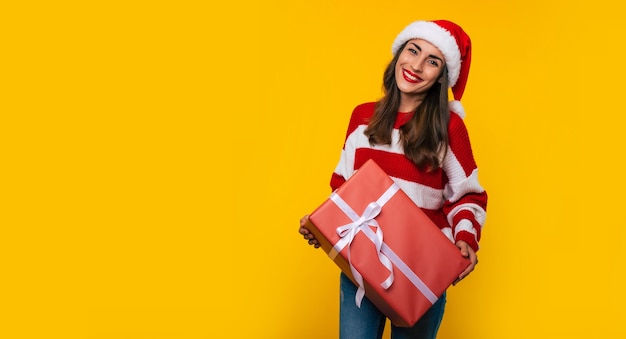 Feche a foto de uma linda mulher sorridente e animada com uma caixa de presente de Natal nas mãos e se divertindo enquanto posa sobre fundo amarelo