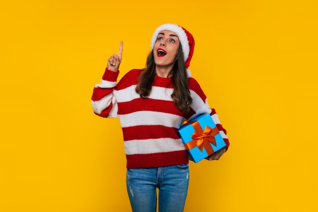 Feche a foto de uma linda mulher sorridente e animada com uma caixa de presente de Natal nas mãos enquanto ela se surpreende apontando para cima em um fundo amarelo