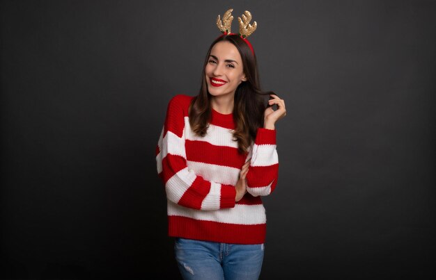 Feche a foto de uma linda mulher sorridente e moderna com chifres de rena de Natal na cabeça enquanto ela posa e se diverte isolada em um fundo cinza
