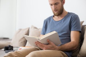 Feche a foto do homem sentado no sofá cinza e lendo cuidadosamente o livro em casa