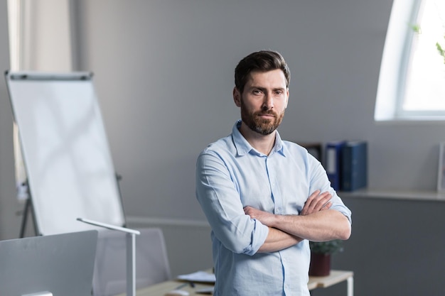 Feche a foto retrato de um jovem no escritório trabalhador de escritório com uma barba em uma camisa leve ele fica com os braços cruzados ele olha para a câmera sorri