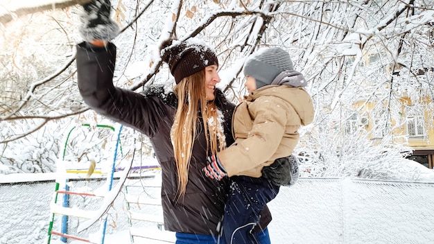 Feche a imagem de uma jovem mulher loira com seu filho fofo na jaqueta e chapéu brincando com uma árvore coberta de neve no parquinho do parque