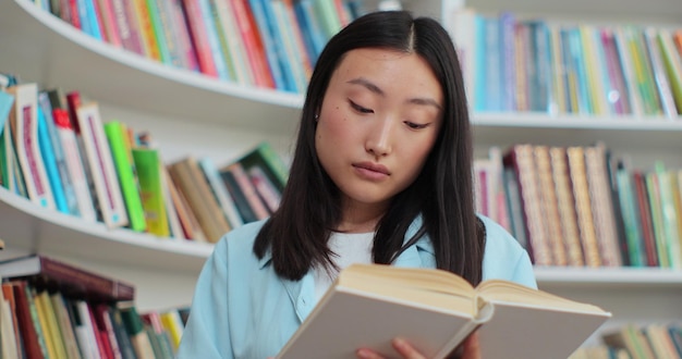 Feche a linda garota asiática lendo livro perto da estante na biblioteca da universidade Conceito de auto-educação