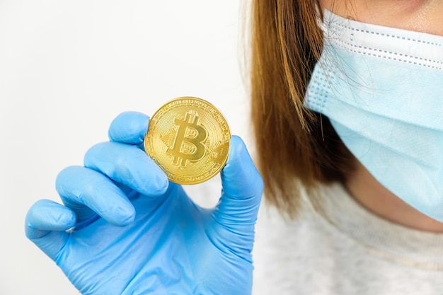 Feche a mão da mulher em luvas de borracha e máscara facial protetora segurando moedas de bitcoin de ouro